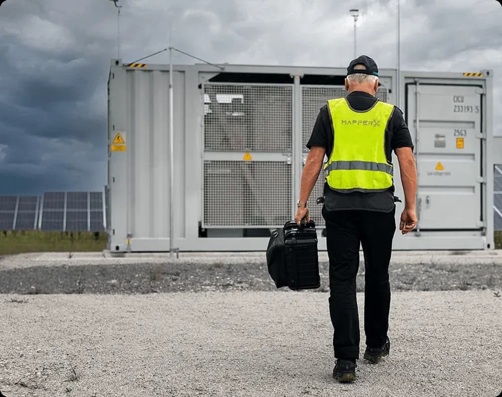 MapperX-technicus loopt met een testkoffer in een zonnepark onder een dreigende lucht naar een container met schakelapparatuur
