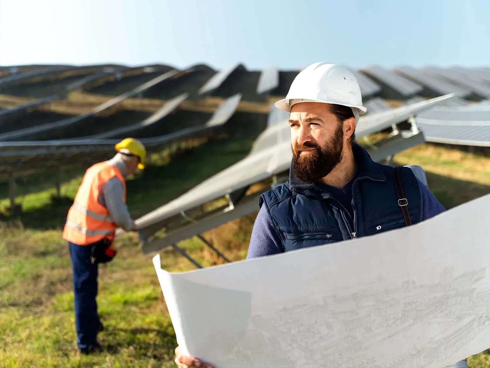 Worker in a hard hat holding a large drawing beside solar panels Worker in a hard hat holding a large drawing beside solar panels